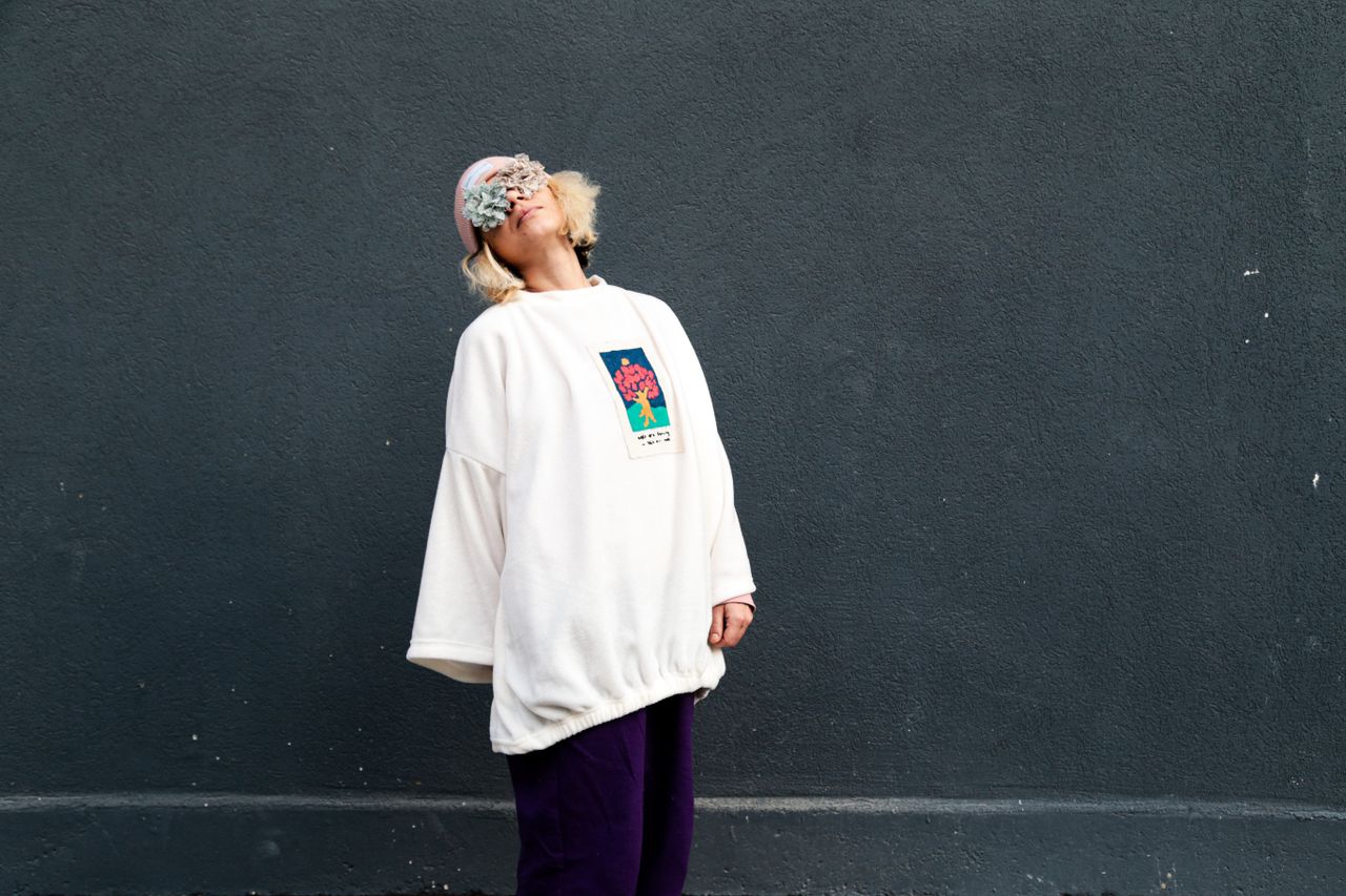 About Person in white sweater and beanie poses artistically against a gray wall.