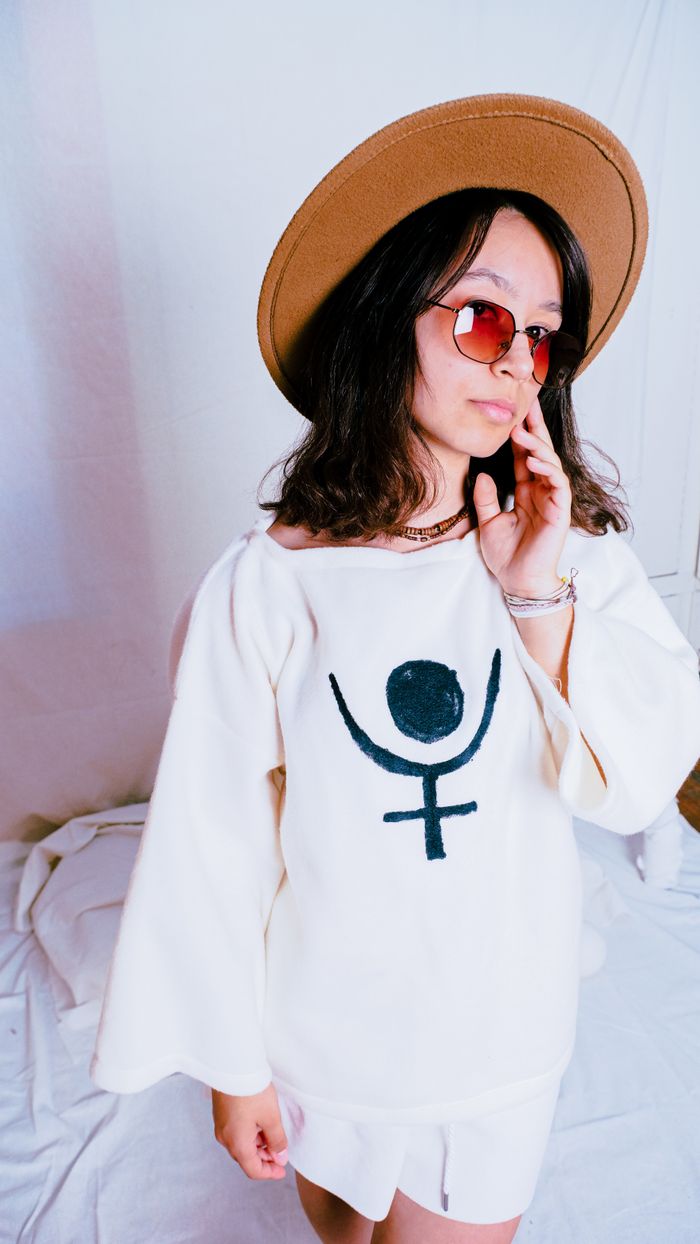 Stylish woman with long hair, wearing a hat and sunglasses, poses in a white outfit with a unique design.