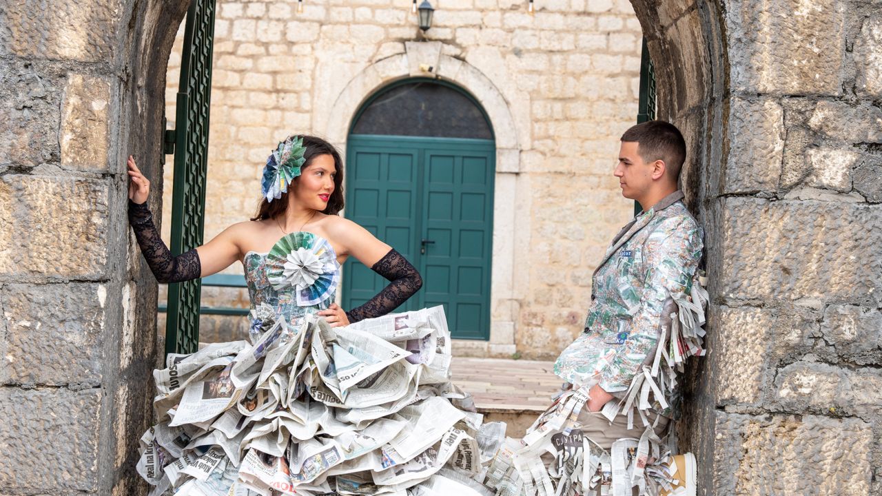 Two people in unique newspaper outfits posed creatively in rustic setting.